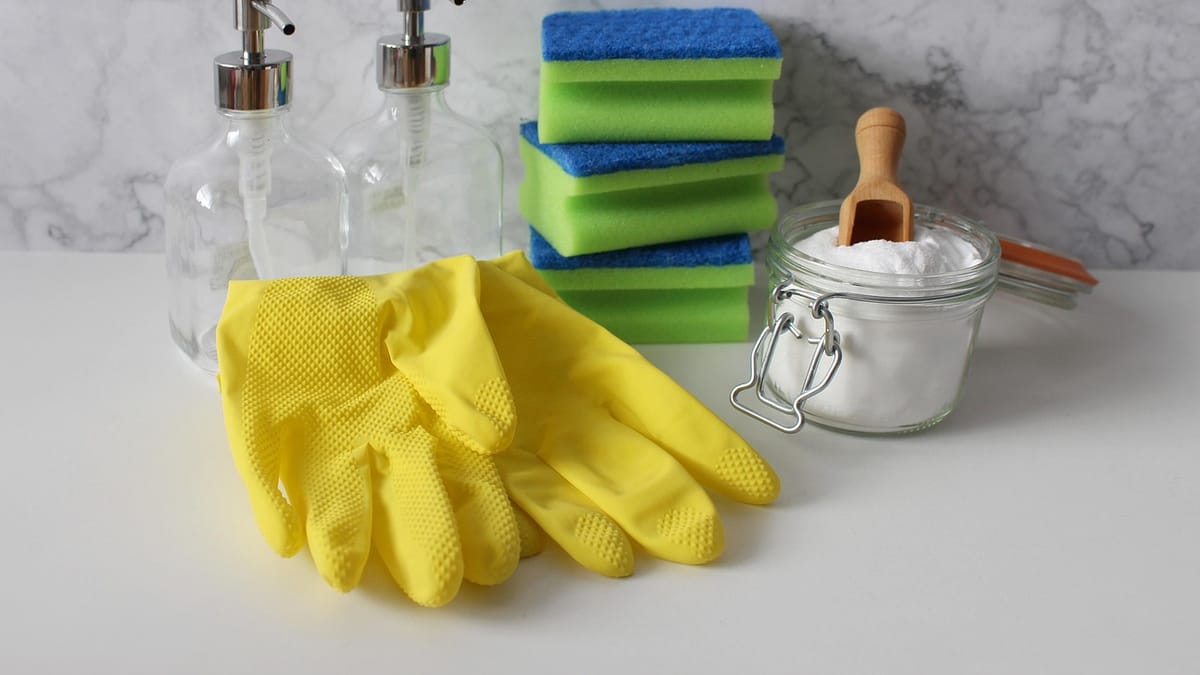 Gants jaunes, éponges empilées, distributeurs en verre et bocal de bicarbonate posés sur un plan de travail blanc